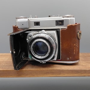 May include: A vintage silver camera with a black lens and a brown leather case. The lens has the text "SYNCHRO" and "Schneider Kreuznach" printed on it. The camera is open, revealing the lens and internal components.