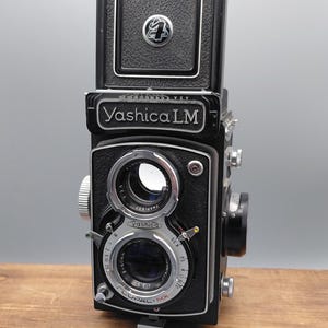 May include: A black Yashica LM twin-lens reflex camera with a top-mounted viewfinder hood. The front features two stacked lenses, a Copal-MX shutter mechanism, and silver metal dials. The camera body has a textured black finish and white text.