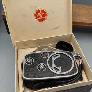 Op de afbeelding: Vintage Bolex Paillard 8mm filmcamera in de originele doos. De camera is zwart met zilveren accenten en een leren handriem. De doos is beige met het Bolex Paillard logo.