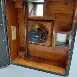 May include: An antique wooden camera with a brass lens and a black leather exterior. The camera has a rectangular shape with a hinged lid and several internal components, including a focusing mechanism and viewfinders. The wood has a warm, reddish-brown tone.
