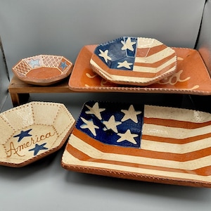 May include: A selection of handcrafted ceramic serving dishes with patriotic designs. The set includes bowls and trays, featuring the American flag, stars, and stripes in red, white, and blue. One bowl has the word "America" written on it.
