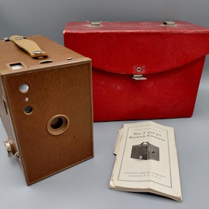 May include: A vintage brown box camera with a tan leather handle and a red case. A booklet titled "Nos. 2 and 2A Brownie Cameras" is next to the camera. The camera has a circular lens and a metal winding knob.