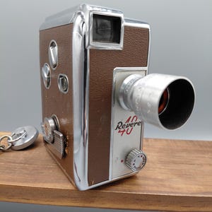 Könnte beinhalten: Vintage Revere 40 Filmkamera mit braunem Kunstledergehäuse und Chromakzenten. Die Kamera hat ein Objektiv, einen Sucher und Bedienknöpfe. Der Name "Revere 40" ist auf der Vorderseite aufgedruckt. Ein silberner Schlüsselanhänger befindet sich links.