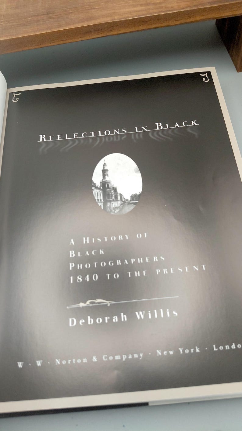 Puede incluir: Portada en blanco y negro del libro "Reflections in Black" de Deborah Willis. Presenta una imagen de un edificio y el texto "A History of Black Photographers 1840 to the Present".