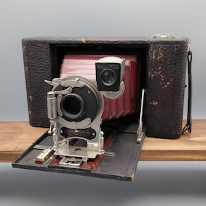 May include: Antique black leather camera with a red bellows and silver-toned lens components. The camera is open, revealing the lens and internal components. The camera sits on a wooden surface.