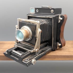 Puede incluir: Una cámara negra de estilo vintage con detalles plateados y un asa de cuero marrón. La cámara presenta una lente con un tinte azul y un diseño de fuelle. La cámara decorativa se encuentra sobre una estantería de madera.