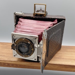 May include: An antique, vintage camera with a red bellows and a silver and brass lens. The camera is open, revealing the bellows. A small, glass-covered display sits on top. The camera is resting on a wooden surface.