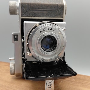May include: Vintage Kodak camera with a silver lens and black leather top. The lens reads "Kodak Ektar f:3.5 50mm Made in U.S.A." A small metal tag with "Kodak" is attached. The camera is on a wooden surface.