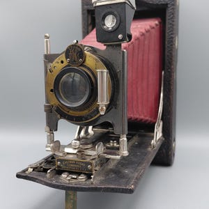 May include: An antique Eastman Kodak camera with a black body, brass lens, and red bellows. The camera has a silver flash attachment and is open, revealing its internal components. The text "Eastman Kodak Company Rochester, N.Y. U.S.A." is visible.