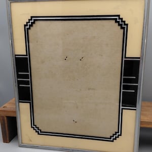 May include: A rectangular picture frame with a metallic silver border. The glass features a cream-coloured background with a black and white geometric border design. The central area is a plain, light-coloured mat board with small dark spots.