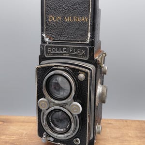 May include: A black Rolleiflex twin-lens reflex camera with a textured leather-like finish. The top viewfinder hood features the name DON MURRAY in gold lettering. The front displays two circular lenses, silver metal dials, and the text ROLLEIFLEX.