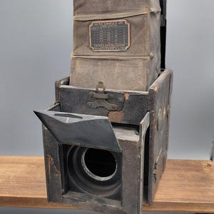 May include: An antique Auto Graflex Jr. camera, with a dark brown leather body and a collapsible bellows. The camera has a lens with an open aperture and a metal plate with text. The camera is placed on a wooden surface.
