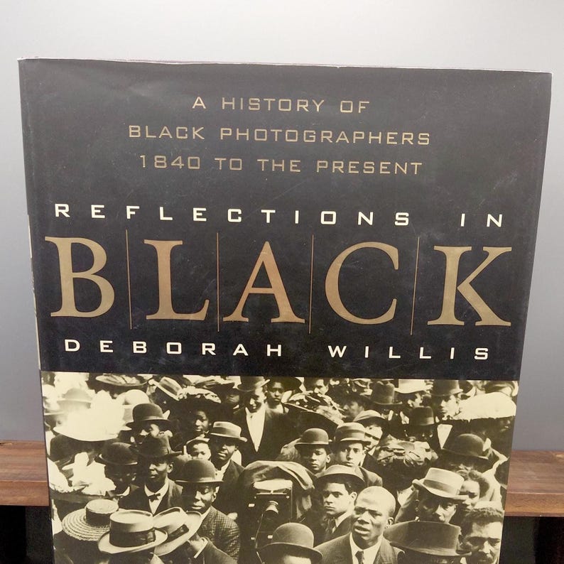 Puede incluir: Un libro titulado "Reflections in Black" de Deborah Willis. La portada presenta un fondo negro con letras doradas y una fotograf&iacute;a en blanco y negro de una multitud. El texto dice "A History of Black Photographers 1840 to the Present."