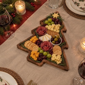 Könnte beinhalten: Ein hölzernes Servierbrett in Form eines Weihnachtsbaums, gefüllt mit einer Auswahl an Lebensmitteln, darunter Salamiröschen, gewürfelter Cheddar-Käse, grüne Trauben, dunkle Trauben, runde Cracker, Brie-Ecken und eine kleine Schale mit Oliven.