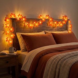 Peut inclure: Une guirlande lumineuse LED composée de feuilles d'érable artificielles dans des tons orange, rouge et jaune. La guirlande illuminée est drapée sur le dessus d'une tête de lit en bois, diffusant une lueur chaude sur le lit et la literie.