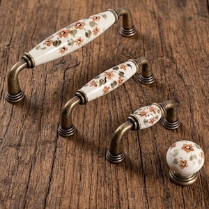 Peut inclure: Ensemble de quincaillerie de meubles de style ancien, comprenant trois poignées et un bouton. Les poignées et le bouton présentent une surface en céramique blanche ornée d'un motif floral brun et orange. Finition bronze.