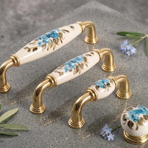 May include: Cabinet hardware set with cream porcelain and blue floral designs. The handles and knob have a gold-coloured metal base. The hardware is designed for use on cabinets and drawers.