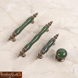 May include: A set of green porcelain cabinet pulls and a knob with antique brass hardware. The pulls have a curved, elongated design, while the knob is round. The hardware has a detailed, ornate design. The items are displayed on a marble surface.