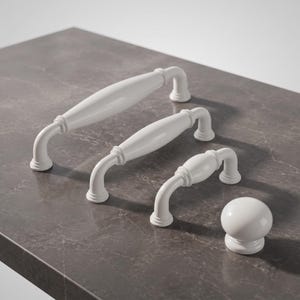 May include: White cabinet hardware set, including two pull handles and one knob. The handles have a curved design with a central, elongated section. The knob is spherical. All pieces are white and sit on a dark gray surface.