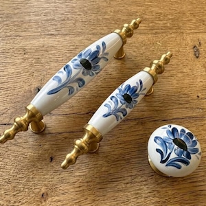 Puede incluir: Dos tiradores de armario florales blancos y azules y un pomo a juego con herrajes de latón. Los tiradores de cerámica tienen un diseño floral azul pintado. El pomo tiene un diseño similar. Los herrajes son dorados.