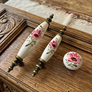 Puede incluir: Dos tiradores de armario de cerámica blanca y un pomo con diseño floral. Los tiradores tienen una base dorada y negra y están pintados con flores rosas y hojas verdes. El pomo tiene un diseño floral a juego.