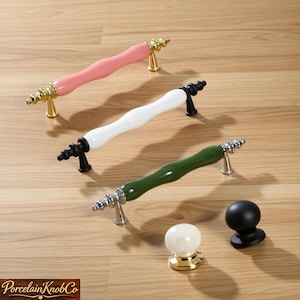 May include: Three cabinet pulls with a pink, white, and green finish, and two cabinet knobs. The pulls have decorative metal ends in gold and silver. The knobs are black and white with gold accents. The items are on a wooden surface.
