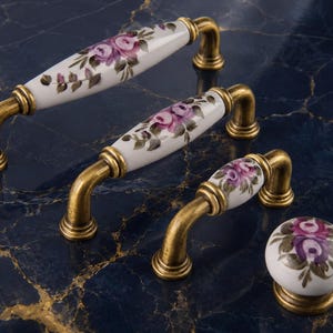May include: A set of cabinet hardware featuring white porcelain handles and a knob, adorned with hand-painted purple rose designs. The hardware has a brass finish and includes two sizes of handles and a knob.