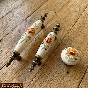 Puede incluir: Juego de herrajes de porcelana color crema para armarios con diseño floral. El juego incluye dos tiradores y un pomo, todos con una flor naranja y marrón pintada a mano con hojas verdes. Los herrajes tienen detalles metálicos color latón antiguo.