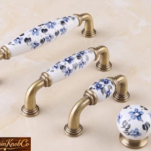 May include: A set of cabinet hardware featuring white porcelain handles and a knob. The porcelain is decorated with blue floral patterns. The hardware has a brass finish, and the set includes two handles and one knob.
