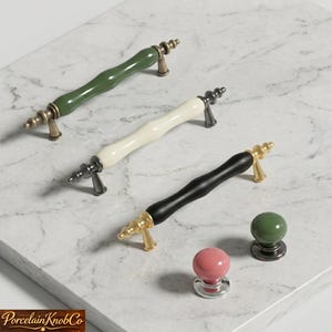 May include: Three cabinet pulls and two knobs in various colors. The pulls have a curved design with brass or silver-toned hardware. Colors include green, white, and black. The knobs are pink and green.
