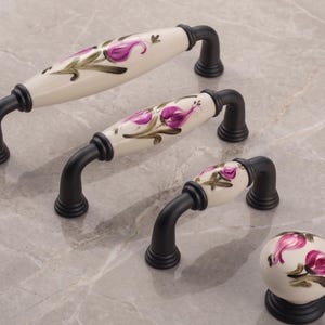 May include: A set of cabinet hardware featuring black metal handles and a knob. The handles have a cream-colored ceramic center with a purple floral design. The knob has a matching floral design and sits on a black base.