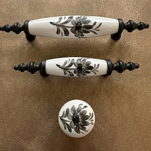 May include: A set of cabinet hardware featuring two white and black floral design handles and a matching knob. The handles have a black metal base and the knob is round. The floral design is in shades of grey and black.