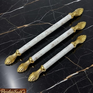 Peut inclure: Quatre poignées de meubles blanches et dorées avec une finition vieillie. Les poignées ont un centre blanc avec des accents dorés aux extrémités. Les extrémités ont un design en forme de feuille. Les poignées sont disposées en diagonale sur une surface en marbre noir.