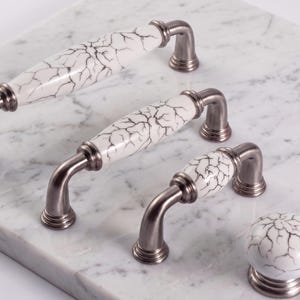 May include: Cabinet hardware set featuring three pull handles and one knob. The handles have a white ceramic center with a black crackle pattern and are paired with brushed nickel hardware. The knob has a matching white ceramic sphere with a crackle design.