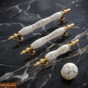 May include: A set of white porcelain cabinet pulls and a knob with gold-colored hardware. The pulls have a curved design with gold filigree patterns. The knob is round with a matching gold design. The hardware is set against a black marble surface.