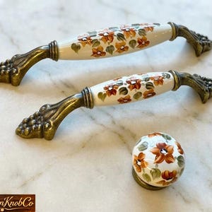 May include: Two white porcelain cabinet pulls and a knob with floral designs in orange, brown, and green. The pulls have bronze-colored grape cluster accents. The Porcelain Knob Co. logo is at the bottom.