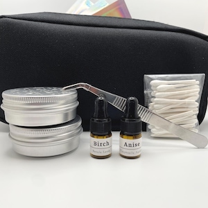 May include: A black zippered pouch containing two round silver metal tins, two small amber glass dropper bottles labeled Birch and Anise, a pair of silver metal tweezers, and a clear plastic bag filled with white cotton swabs.