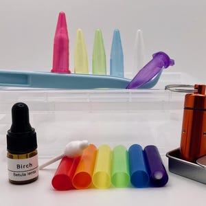 May include: A collection of laboratory supplies including a clear plastic storage box, a light blue plastic tool, a small amber glass bottle labeled Birch Betula lenta, a metallic orange cylindrical container, and various colorful plastic tubes and caps.