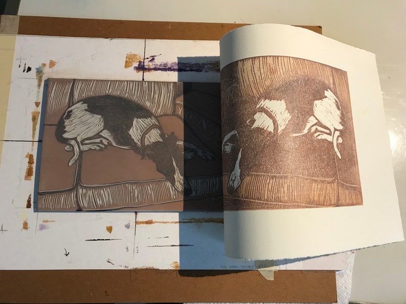K&ouml;nnte beinhalten: Eine Nahaufnahme eines braunen und wei&szlig;en Drucks eines Hundes, der auf einem Sofa liegt. Der Druck ist auf einem wei&szlig;en Blatt Papier und wird auf eine braune Oberfl&auml;che gelegt.