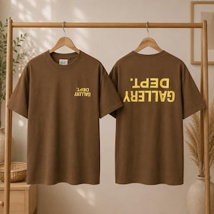 May include: Two brown short-sleeve t-shirts hanging on wooden hangers. The left shirt features a small, inverted yellow text reading GALLERY DEPT. on the chest. The right shirt displays the same text in a larger, inverted yellow font on the back.