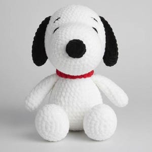 Könnte beinhalten: Handgefertigtes, weißes Häkelhund-Plüschtier mit schwarzen Ohren und einer schwarzen Nase. Der Hund hat ein rotes Halsband und sitzt mit ausgestreckten Armen und Beinen. Das Plüschtier ist aus weichem, strukturiertem Garn.