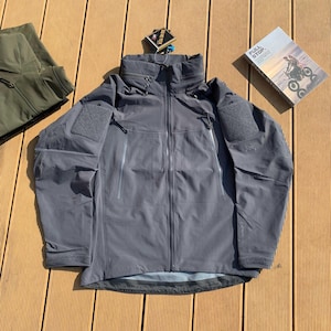 May include: A grey tactical jacket with a high collar and multiple pockets. The jacket has a full-length zip and patches on the sleeves. A tag with the text "GORE-TEX" is attached. A book with the text "FULL STOP" is also visible.