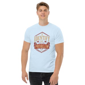 Może przedstawiać: Jasnoniebieski t-shirt z krótkim rękawem z grafiką w czerwono-złotej sześciokątnej ramce. Napis I IDENTIFY AS A PROBLEM wykonano pogrubioną, postarzaną czcionką, a u dołu grafiki umieszczono dwie skrzyżowane pałeczki perkusyjne.