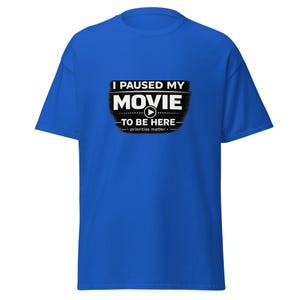 Op de afbeelding: Koningsblauw T-shirt met de tekst "I PAUSED MY MOVIE TO BE HERE - priorities matter." in wit en zwart, met een afspeelknop. Het shirt is een casual stijl met korte mouwen.