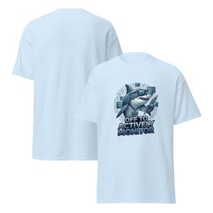 May include: Light blue t-shirt featuring a cartoon shark wearing sunglasses and holding a pen. The shirt has the text "OFF TO ACTIVELY MONITOR" in a blue font. The back of the shirt is plain light blue.