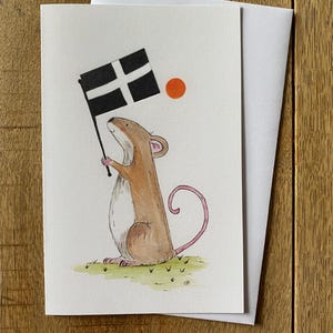 May include: A watercolor greeting card featuring a brown mouse holding a black and white flag with a white cross. The mouse is standing on green grass with a small orange circle above the flag. A white envelope is behind the card.