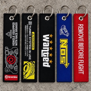 Op de afbeelding: Vijf rechthoekige stoffen sleutelhangers met metalen ringen. De eerste is zwart met witte tekst en een rood Brembo-logo. De tweede is zwart met gele en witte tekst. De derde is zwart met witte tekst en gele sterren. De vierde is blauw met gele NOS-tekst. De vijfde is rood met witte tekst.