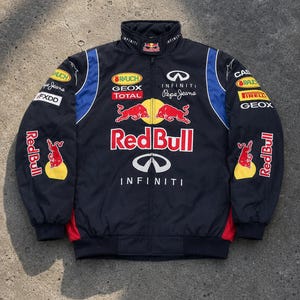 Könnte beinhalten: Eine schwarze Jacke mit Reißverschluss und den Logos von Red Bull, Infiniti, Pepe Jeans, Rauch, Geox, Total, Pirelli und FXDD. Die Jacke hat blaue und rote Seiteneinsätze, einen hohen Kragen mit Red Bull-Logo sowie lange Ärmel mit gerippten Bündchen.