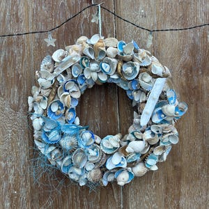 Op de afbeelding: Een ronde krans opgebouwd uit een dichte verzameling van diverse schelpen in tinten wit, bruin en blauw. Het ontwerp bevat kleine witte parels, stukjes blauw net en een lange witte schelp, alles opgehangen aan een metalen haak.