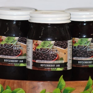 May include: Three jars of Boysenberry Jam from Das Am Shoppe, each with a white lid and a label featuring a berry illustration. The jars are displayed on a wooden tray with green foliage in the foreground.